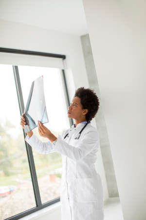 Female African American doctor wearing white coat with stethoscope standing by the window in the office and looking x-ray imageの写真素材