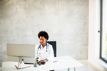 Female African American doctor wearing white coat with stethoscope sitting behind desk in the officeの写真素材