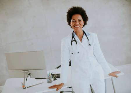 Female African American doctor wearing white coat with stethoscope standing by desk in the officeの写真素材