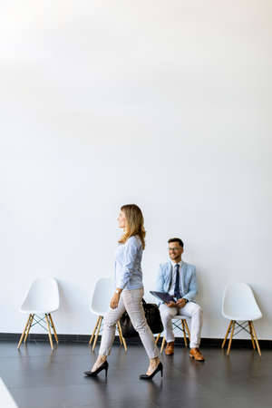 Businesswoman with suitcase passing by young man sitting at chair in the waiting room with a folder...
