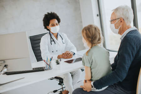 Grandfather with protective facial masks with cute granddaughter at black female doctor