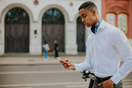 Handsome young African American using mobile phone while standing with electric scooter on a streetの写真素材