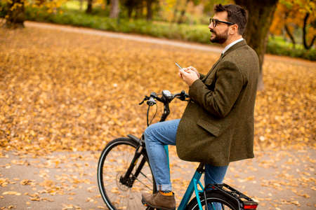Handsome young man using mobile phone on electric bicycle at the autumn park