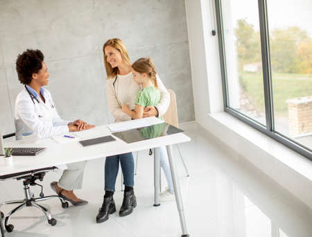 Cute little girl with her mother at the pediatrician examination by African american female doctorの写真素材