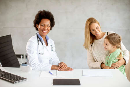 Cute little girl with her mother at the pediatrician examination by African american female doctorの写真素材