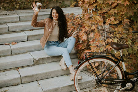 Pretty young woman with mobile phone drink coffee to go at the stairs by the bicycle on autumn day