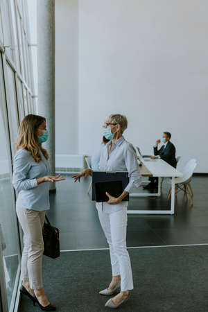 Two businesswomen, mature and young one, talking in the office and wearing mask as a virus...