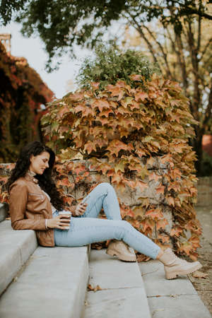 Pretty young woman with mobile phone drink coffee to go at the outdoor stairs on autumn dayの写真素材