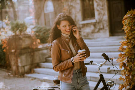 Pretty young woman with mobile phone drink coffee to go by the bicycle on autumn dayの写真素材