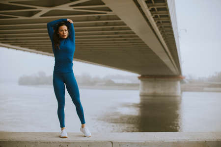 Pretty young woman in blue track suit stretching before workout by the river at autumn morningの写真素材