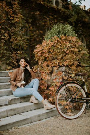 Pretty young woman with mobile phone drink coffee to go at the stairs by the bicycle on autumn dayの写真素材