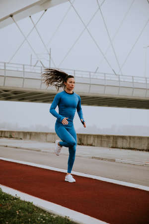 Pretty young woman in blue track suit running by the river at autumn morningの写真素材