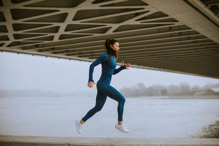 Pretty young woman in blue track suit running by the river at autumn morningの写真素材