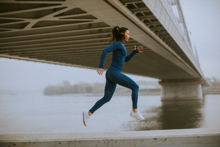 Pretty young woman in blue track suit running by the river at autumn morningの写真素材