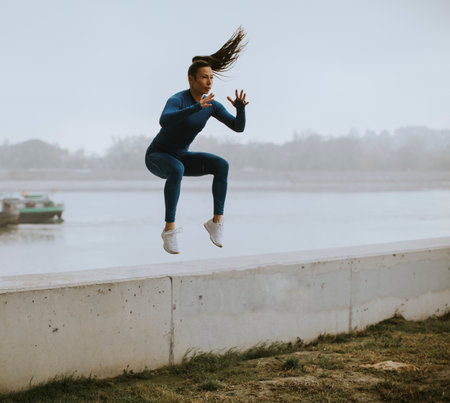 Pretty young woman in blue track suit taking jump by the river at autumn morningの写真素材