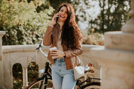 Pretty young woman with mobile phone drink coffee to go by the bicycle on autumn dayの写真素材