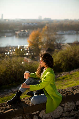 Pretty young curly hair woman enjoying autumn sun while sitting by the river and drinking takeaway coffeeの写真素材