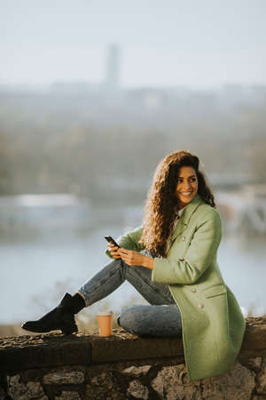 Pretty young curly hair woman using smartphone while sitting by the river and drinking takeaway coffeeの写真素材