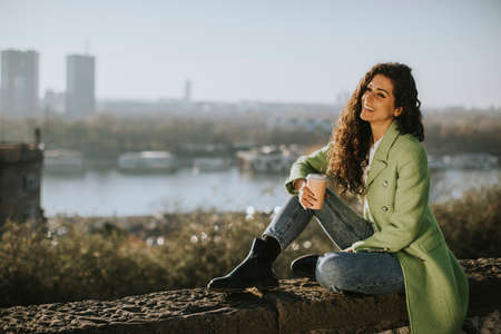 Pretty young curly hair woman enjoying autumn sun while sitting by the river and drinking takeaway coffeeの写真素材