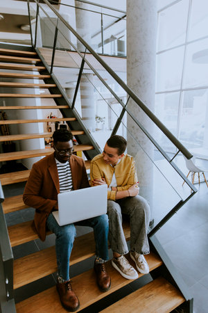 Handsome young African American business man and short hair woman working on laptop while sitting at office stairsの写真素材