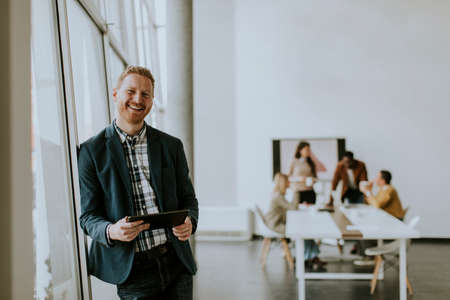 Young handsome businessman standing with digital tablet in front of his team at the office