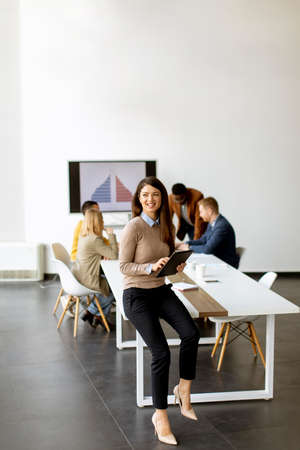 Cute young business woman standing in the office and using digital tablet in front of her team
