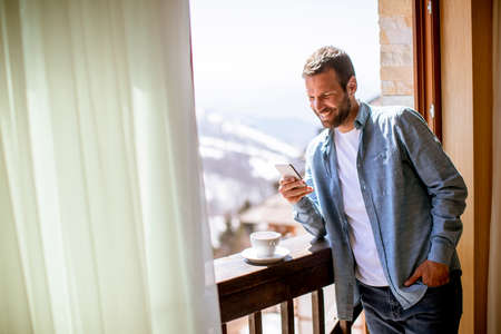 Handsome young man using mobile phone with cup of hot tea at the winter window