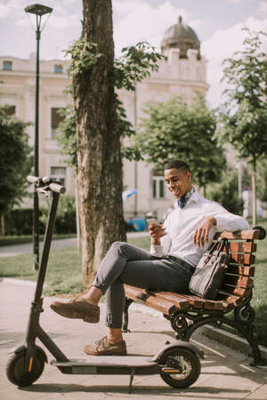 Handsome young African American using mobile phone while sitting on the bench by electric scooterの写真素材