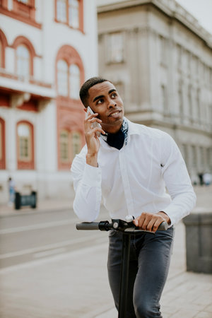 Handsome young African American using mobile phone while standing with electric scooter on a streetの写真素材