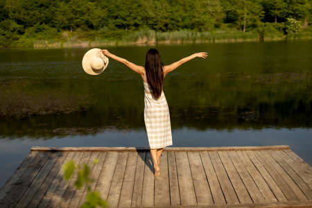 Happy young woman standing on the wooden pier at the calm lake on a hot summer dayの写真素材