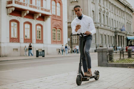 Handsome young African American using mobile phone while standing with electric scooter on a streetの写真素材