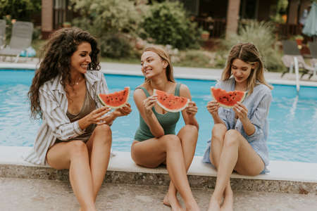 Three cute young women sitting on by the swimming pool and eating watermelon in the house backyard