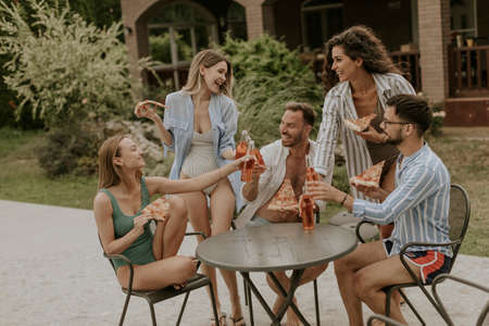 Group of happy young people cheering with cider and eating pizza by the pool in the garden