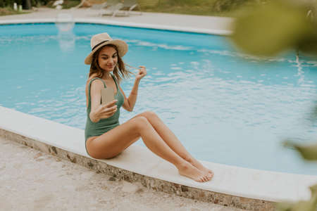 Cute young woman sitting by the swimming pool and taking selfie photo with  mobile phone in the...