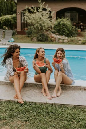 Three cute young women sitting on by the swimming pool and eating watermelon in the house backyard