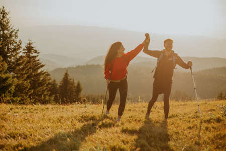 Smiling young couple walking with backpacks over green hillsの写真素材