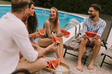 Group of young people sitting by the swimming pool and eating watermelon in the house backyard