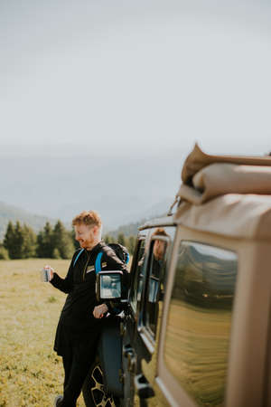 Handsome young man relaxing and drinking coffee by the terrain vehicle hood at countryside