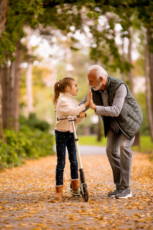 Senior man teaching his granddaughter how to ride kick scooter in the autumn parkの写真素材