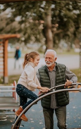 Handsome grandfather spending time with his granddaughter in park playground on autumn day
