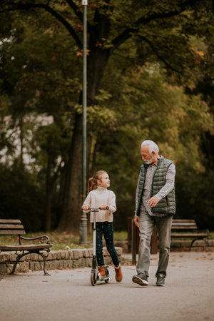 Senior man teaching his granddaughter how to ride kick scooter in the autumn parkの写真素材