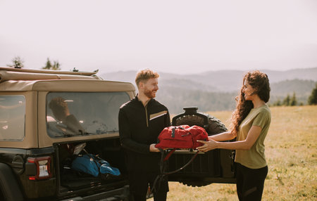 Smiling young couple preparing hiking adventure with backpacks by terrain vehicleの写真素材