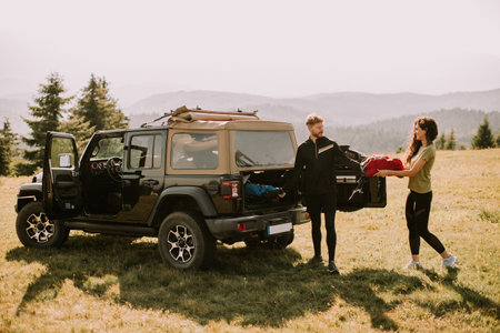 Smiling young couple preparing hiking adventure with backpacks by terrain vehicle