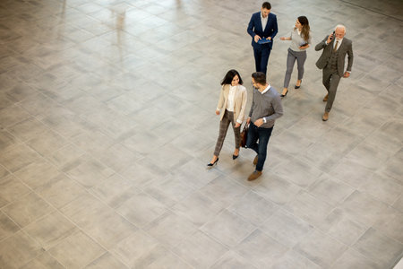 Group of young and senior business people are walking in an office hallway, captured in an aerial view. They are dressed in formal attire, walking with purpose and intent and discussing amongst themselves and interacting with colleaguesの写真素材
