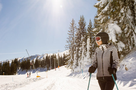 Young woman enjoying winter day of skiing on the snow covered slopes, surrounded by tall trees and dressed for cold temperaturesの写真素材