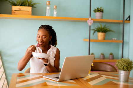 Pretty young black woman having a healthy breakfast while working on laptop in the cafeの写真素材