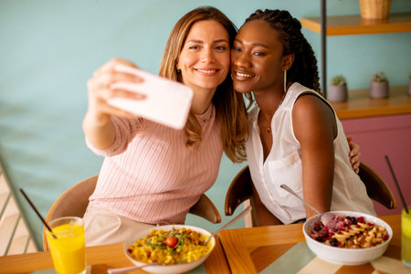 Two pretty young women, caucasian and black one, taking selfie with mobile phone in the cafeの写真素材
