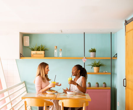 Two pretty young black and caucasian woman having good time, drinking fresh juices and having healthy breakfast in the cafeの写真素材