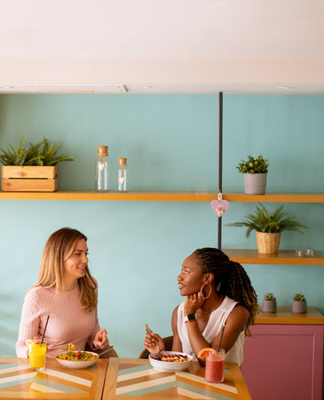 Two pretty young black and caucasian woman having good time, drinking fresh juices and having healthy breakfast in the cafeの写真素材