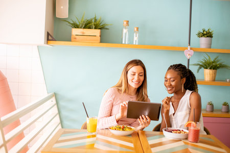 Two pretty young black and caucasian woman having good time, drinking fresh juices and having healthy breakfast in the cafeの写真素材